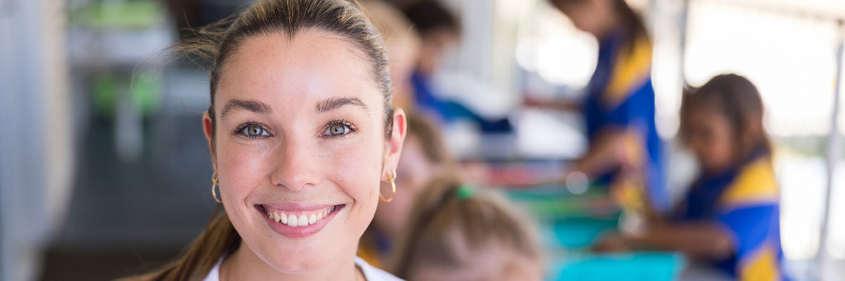 Smiling female teacher in classroom with students in the background.